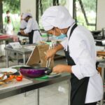 Culinary students in uniforms practice cooking skills in a professional kitchen setting.